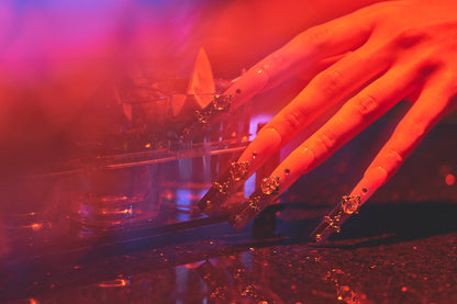 Hand with Divine Plea pink press-on nails holding a tray of tequila shots with lime wedges, captured under dramatic red and purple lighting.