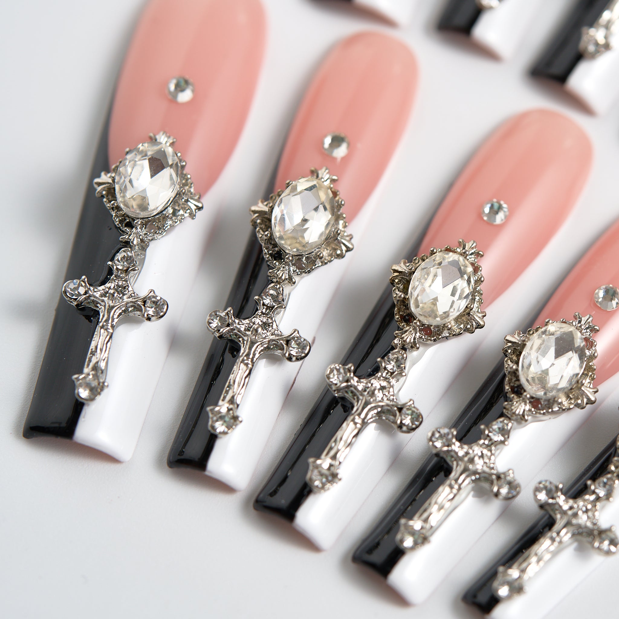 Angled close-up of Divine Plea press-on nails from MiuNail, featuring aligned silver cross charms, large rhinestones, and glossy black and white tips on a nude base over white background.