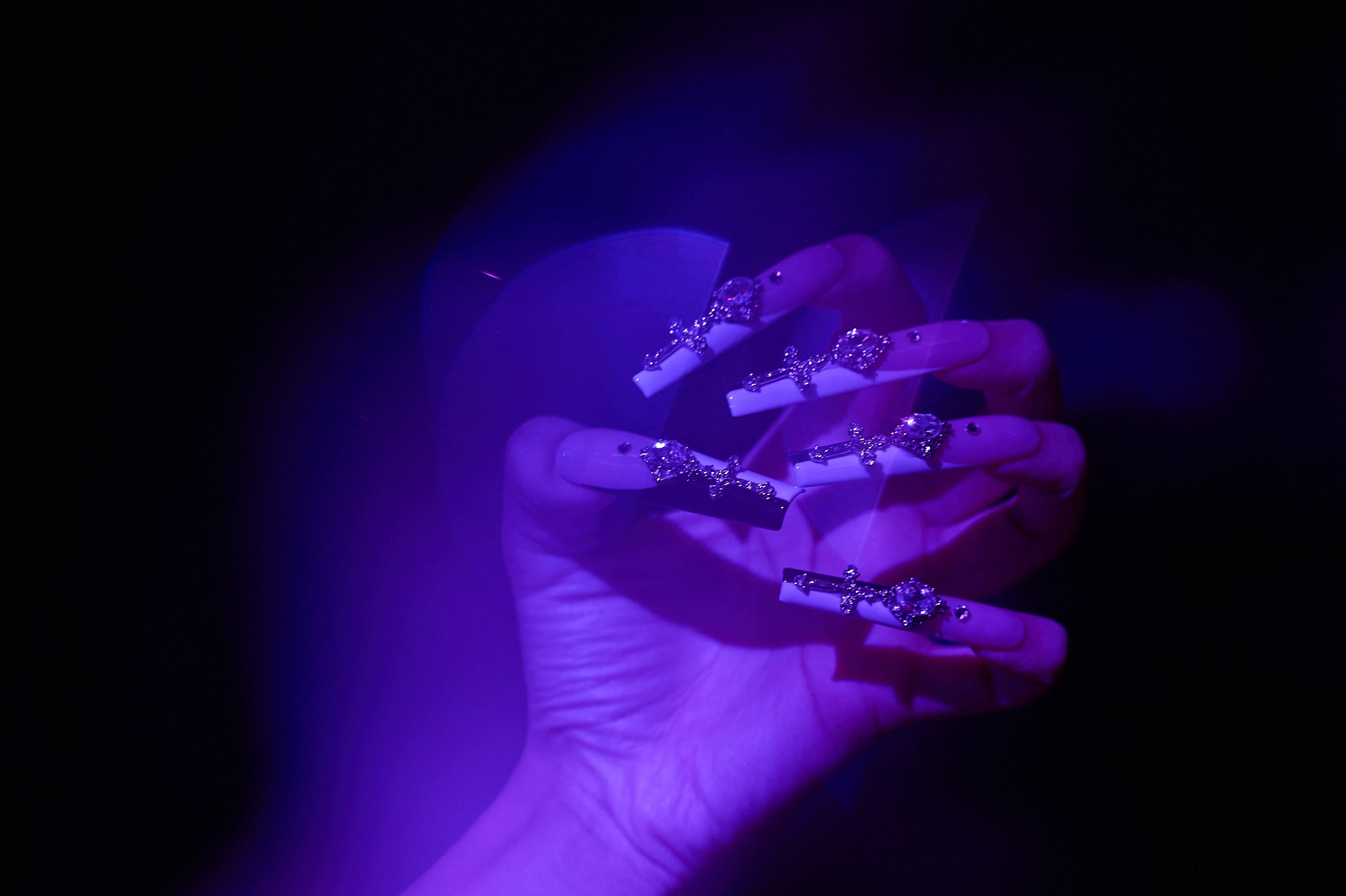 Divine Plea press-on nails with silver rhinestone crosses and crystals, captured under moody purple lighting in a stylized hand pose.