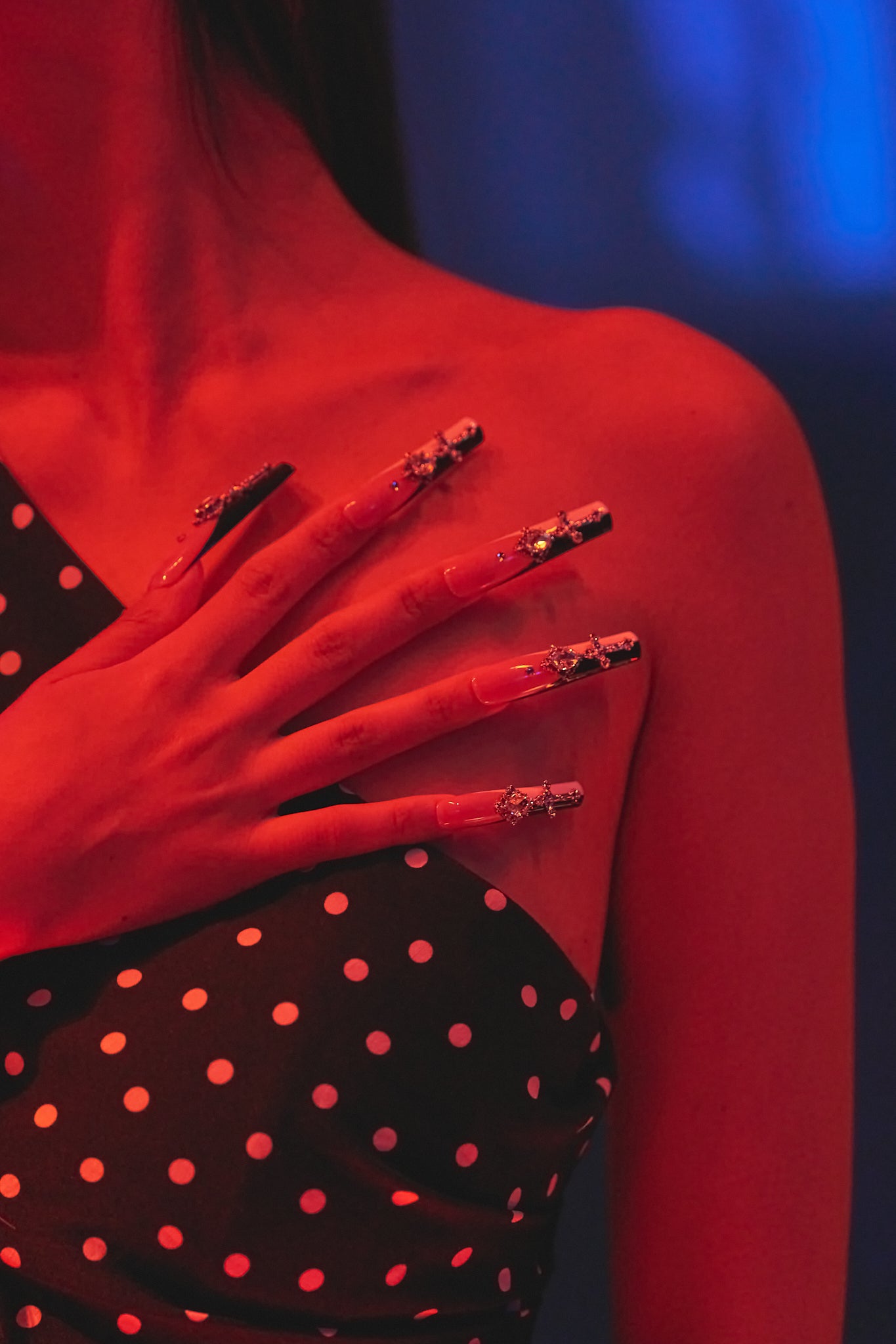 Close-up of a hand wearing MiuNail Divine Plea cross press-on nails, resting on a black polka dot dress under warm red lighting.