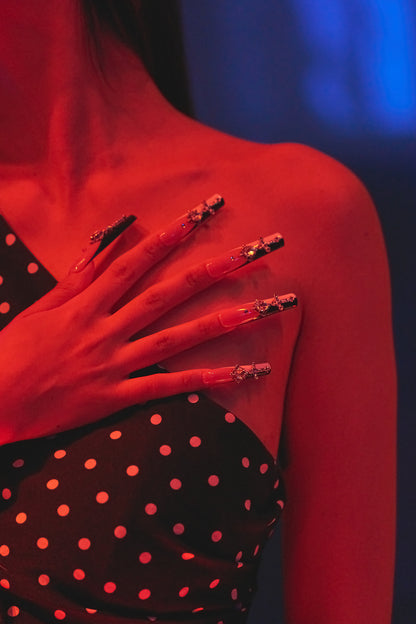 Close-up of a hand wearing MiuNail Divine Plea cross press-on nails, resting on a black polka dot dress under warm red lighting.
