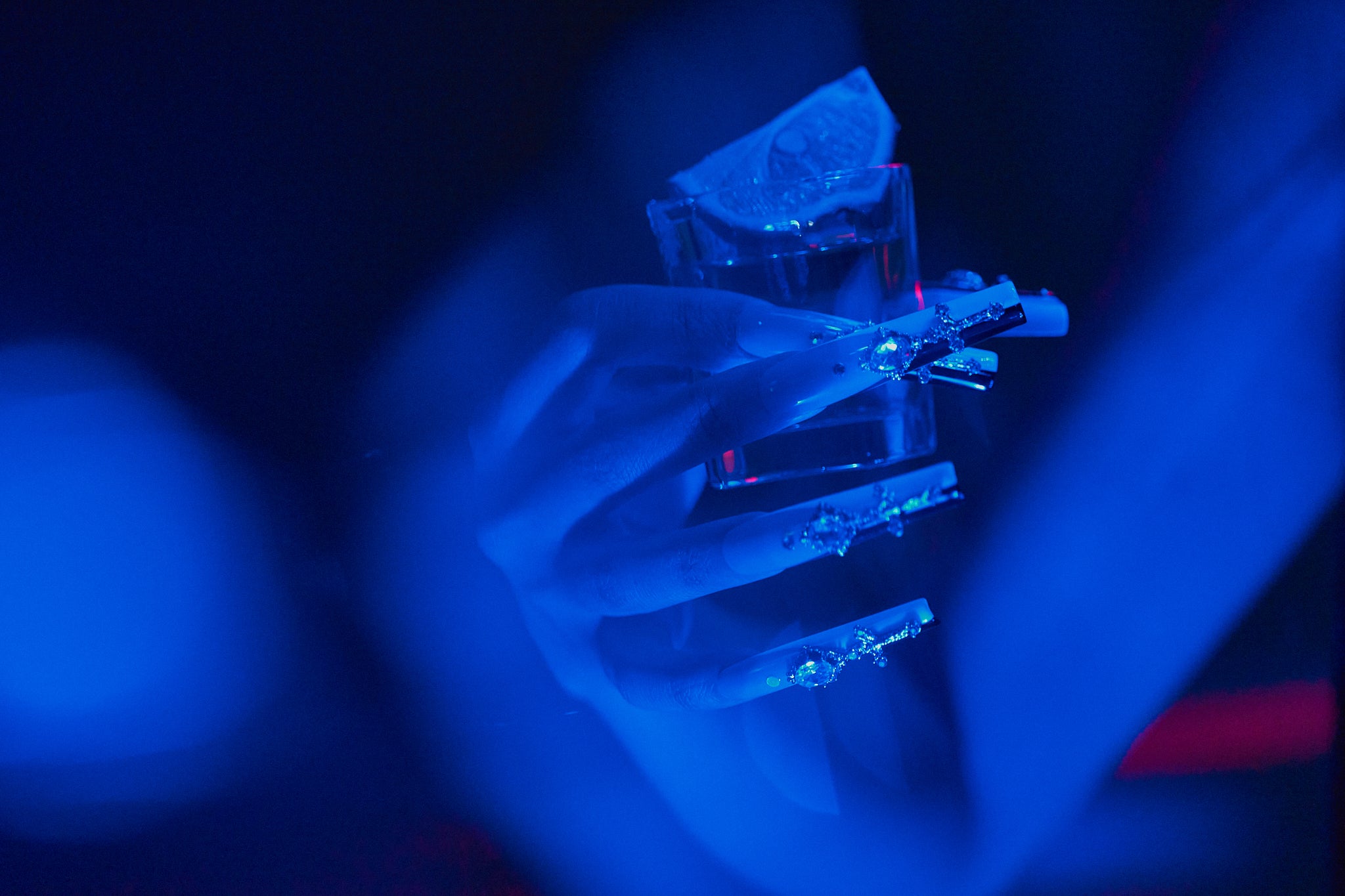Divine Plea press-on nails by MiuNail featuring crystal crosses and rhinestones, shown under deep blue lighting while holding a tequila shot with a lime wedge.