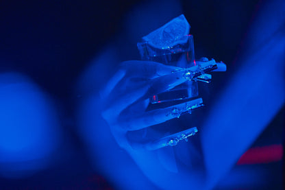 Divine Plea press-on nails by MiuNail featuring crystal crosses and rhinestones, shown under deep blue lighting while holding a tequila shot with a lime wedge.
