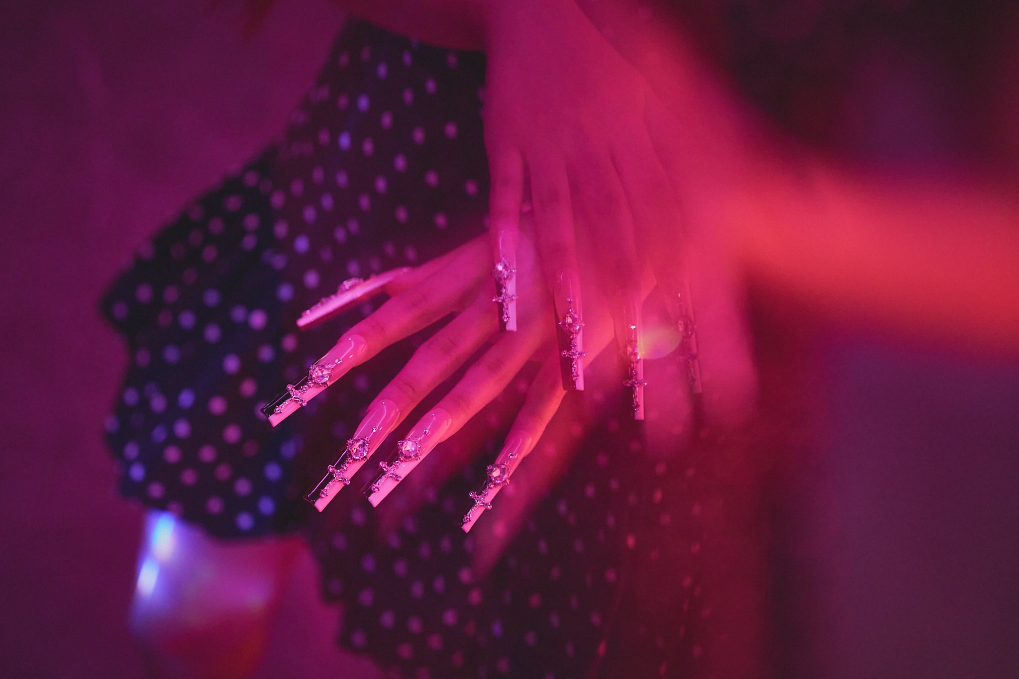 Divine Plea press-on nails by MiuNail featuring rhinestone crosses, shown on a hand resting against a black polka-dot dress under pink ambient lighting.