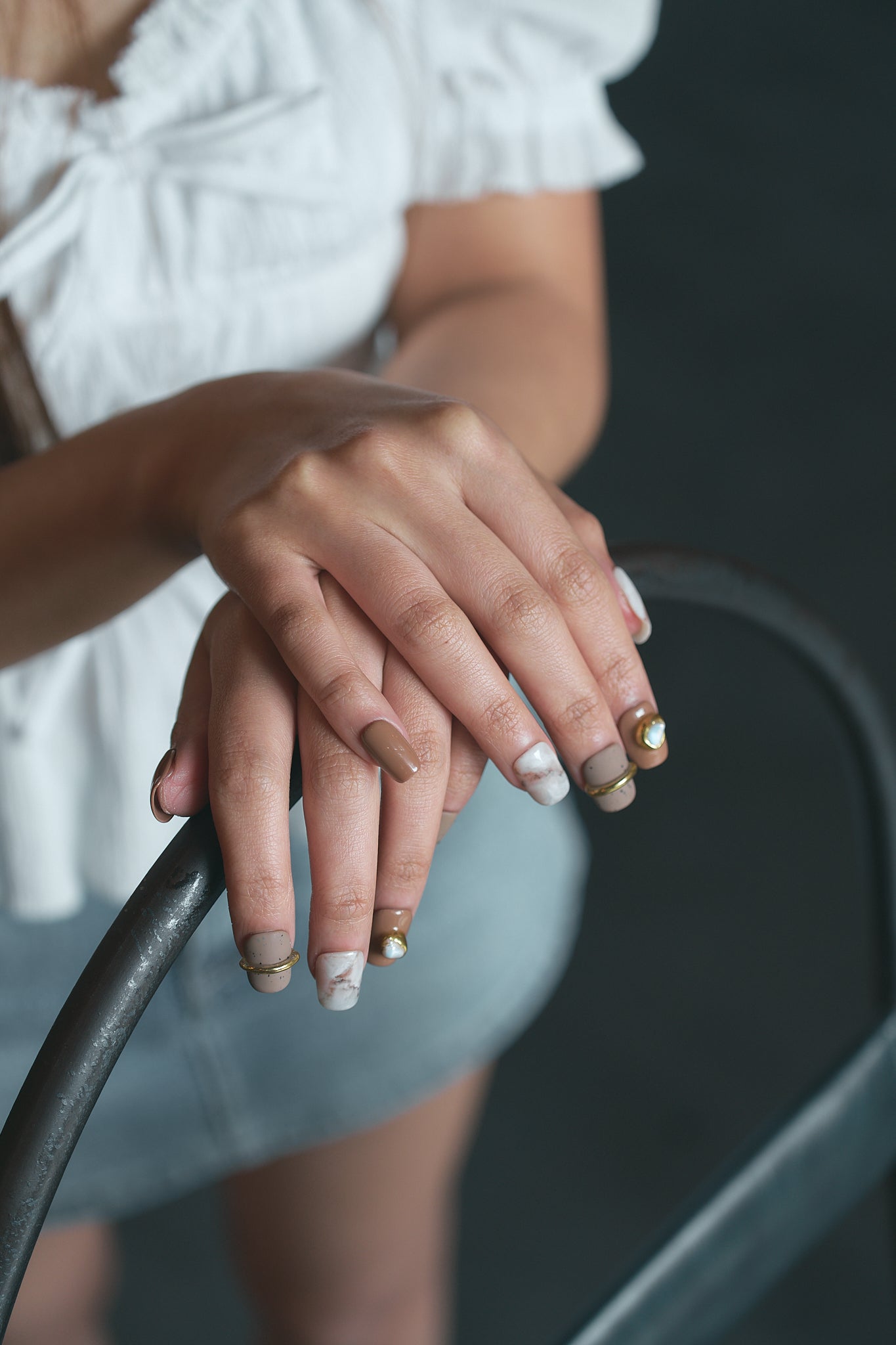 Hands with MiuNail Golden Dune press-on nails in marble and brown with gold accents, resting on a metal bar, person wearing a white top and denim shorts.