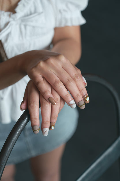 Hands with MiuNail Golden Dune press-on nails in marble and brown with gold accents, resting on a metal bar, person wearing a white top and denim shorts.