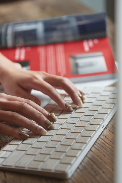 Hands with MiuNail Golden Dune press-on nails in marble, brown, and gold, typing on a white wireless keyboard with a red book and magazine in the background.
