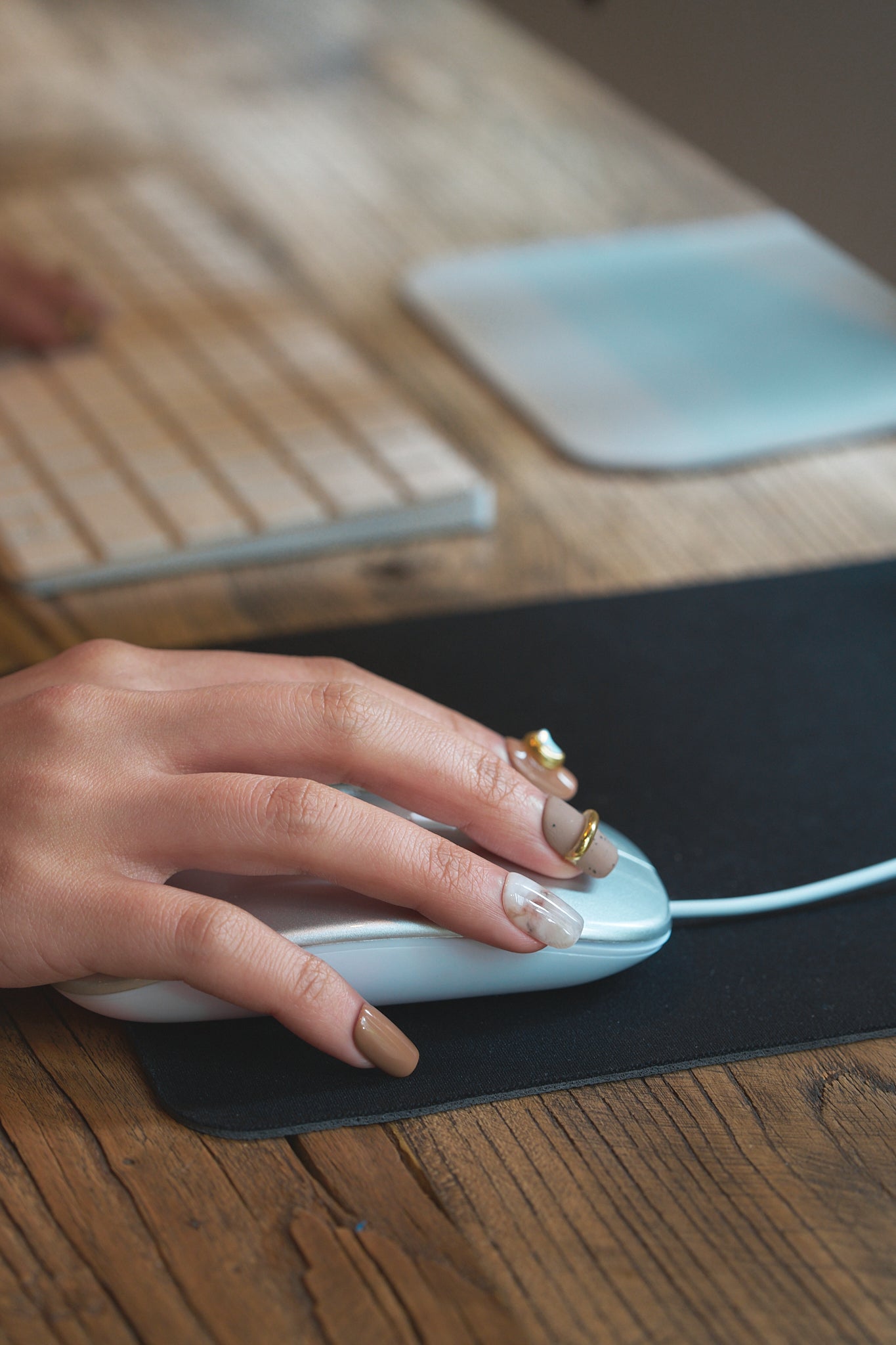 Hand wearing MiuNail Golden Dune press-on nails in marble and brown with gold accents, using a computer mouse on a wooden desk, keyboard in the background.