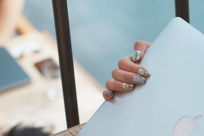 Hand with MiuNail Mist Orbit metallic press-on nails holding a closed laptop outdoors.