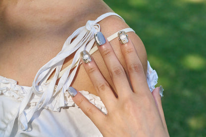 Hand with MiuNail Mist Orbit press-on nails and decorative rings resting on a white lace top, outdoors with green background.
