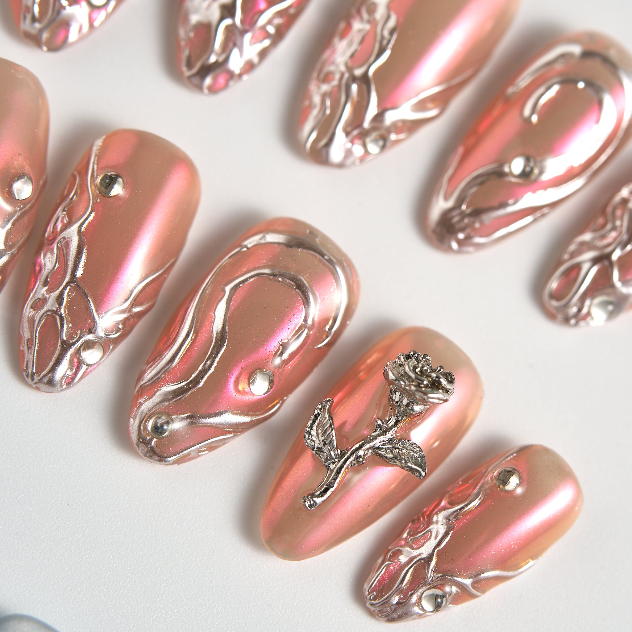 Close-up of MiuNail Molten Coral press-on nails in coral chrome with raised silver detailing and rhinestones, displayed in soft studio lighting.