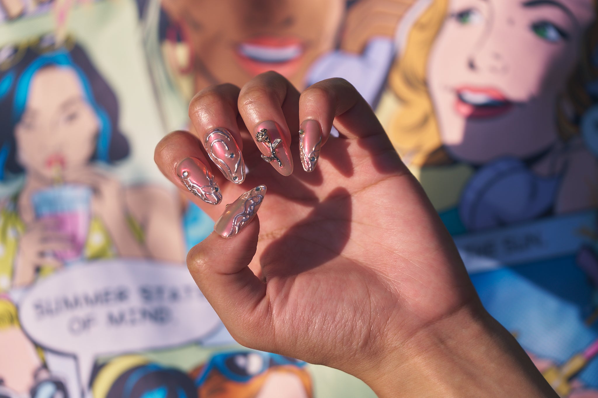 Hand wearing MiuNail Molten Coral press-on nails in coral pink chrome with silver detailing, posed in front of a comic-style mural.