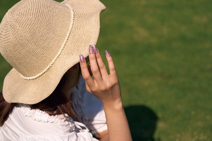 Person wearing MiuNail Molten Coral press-on nails in metallic coral pink with chrome accents, holding a straw hat outdoors in sunlight.
