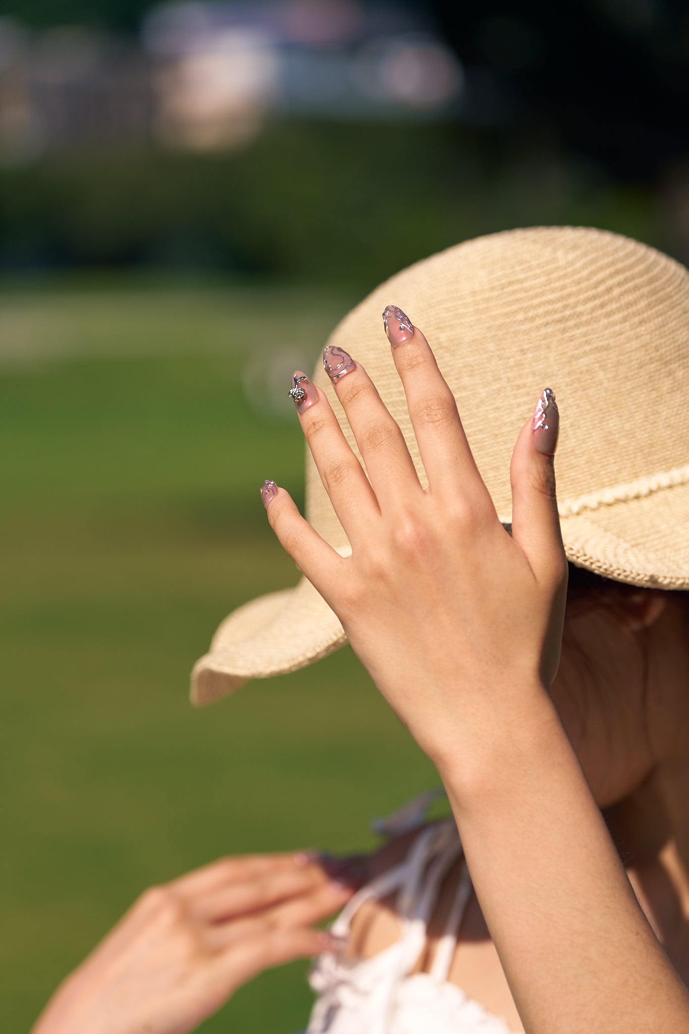 Hand with MiuNail Molten Coral press-on nails in shiny coral chrome, gently gripping a straw hat with green foliage in the background.