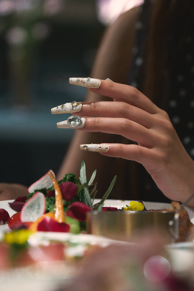 Hand with intricate white and gold embellished nails reaching toward a plate of colorful food, fine detail of MiuNail Ornate Veil set.