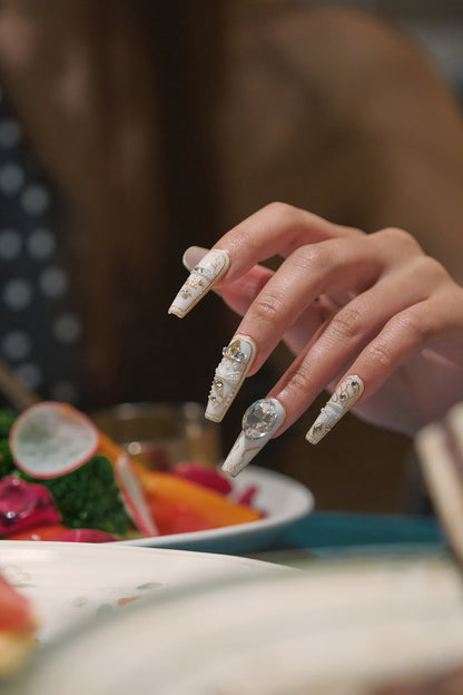 Hand with detailed white and gold nail art resting above a colorful salad at a dinner table.