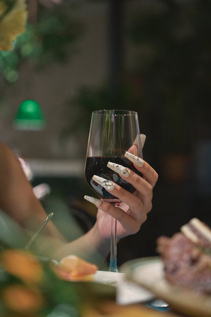 Hand wearing MiuNail Ornate Veil press-on nails with gold and crystal embellishments, elegantly holding a glass of red wine at a dinner table.