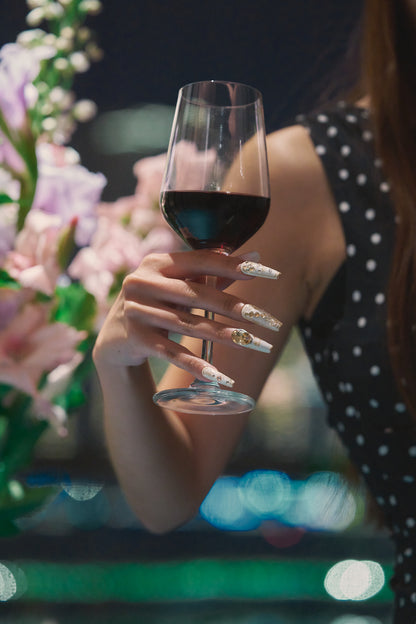 Hand wearing MiuNail Ornate Veil press-on nails with gold and crystal details, holding a glass of red wine with blurred flowers and lights in the background.