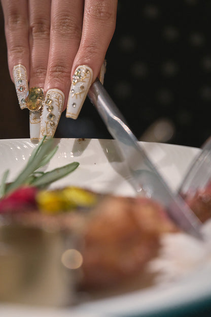 Hand with ornate nail designs using tongs to serve food, focus on nail art details and dining scene.