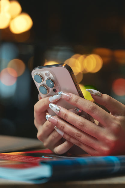Hand with MiuNail “Petal Bloom” floral press-on nails holding a smartphone, with a soft bokeh background.