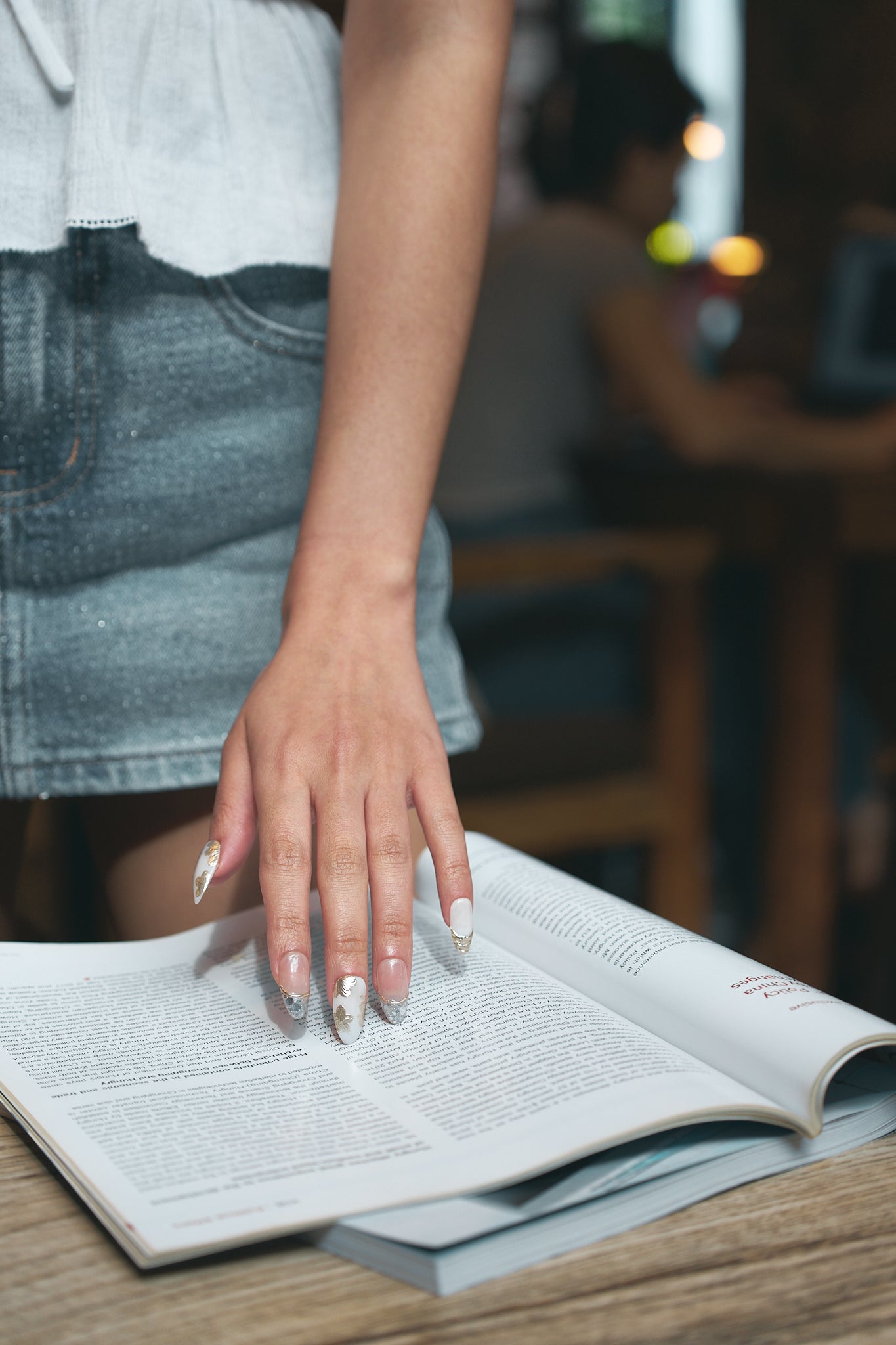 Petal Bloom long almond press-on nails in marble white and gold, hand resting on an open book in a casual setting