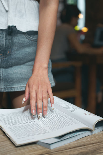 Petal Bloom long almond press-on nails in marble white and gold, hand resting on an open book in a casual setting