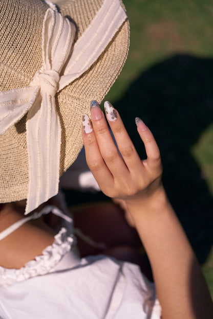 Petal Bloom long almond press-on nails in marble white and gold, hand raised beside straw hat with bow outdoors