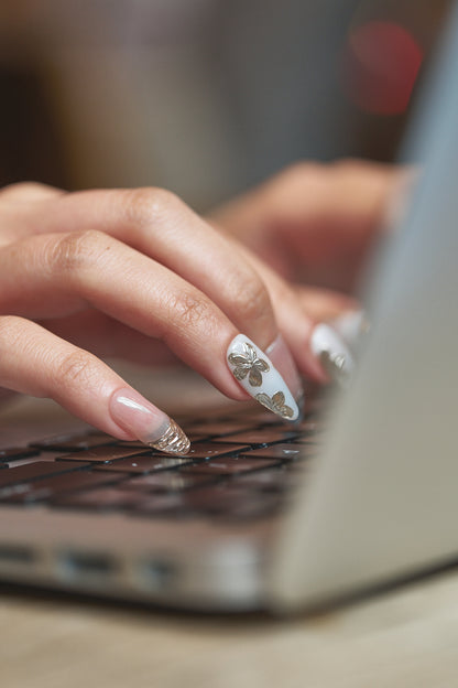Hand with MiuNail “Petal Bloom” short almond press-on nails in glossy nude with white floral accents, typing on a laptop.