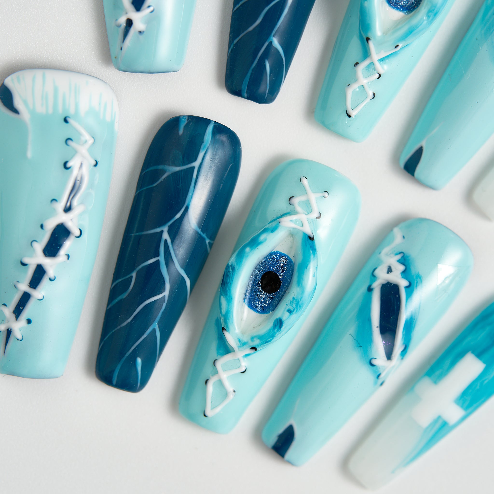 Top-down close-up of MiuNail Siren Hex press-on nails in icy blue and navy with stitched designs, lightning patterns, and a raised eye detail, arranged on a white surface.
