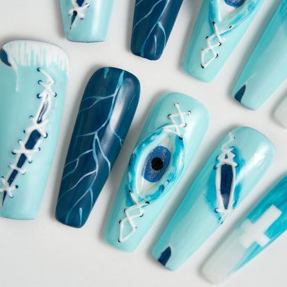 Top-down close-up of MiuNail Siren Hex press-on nails in icy blue and navy with stitched designs, lightning patterns, and a raised eye detail, arranged on a white surface.
