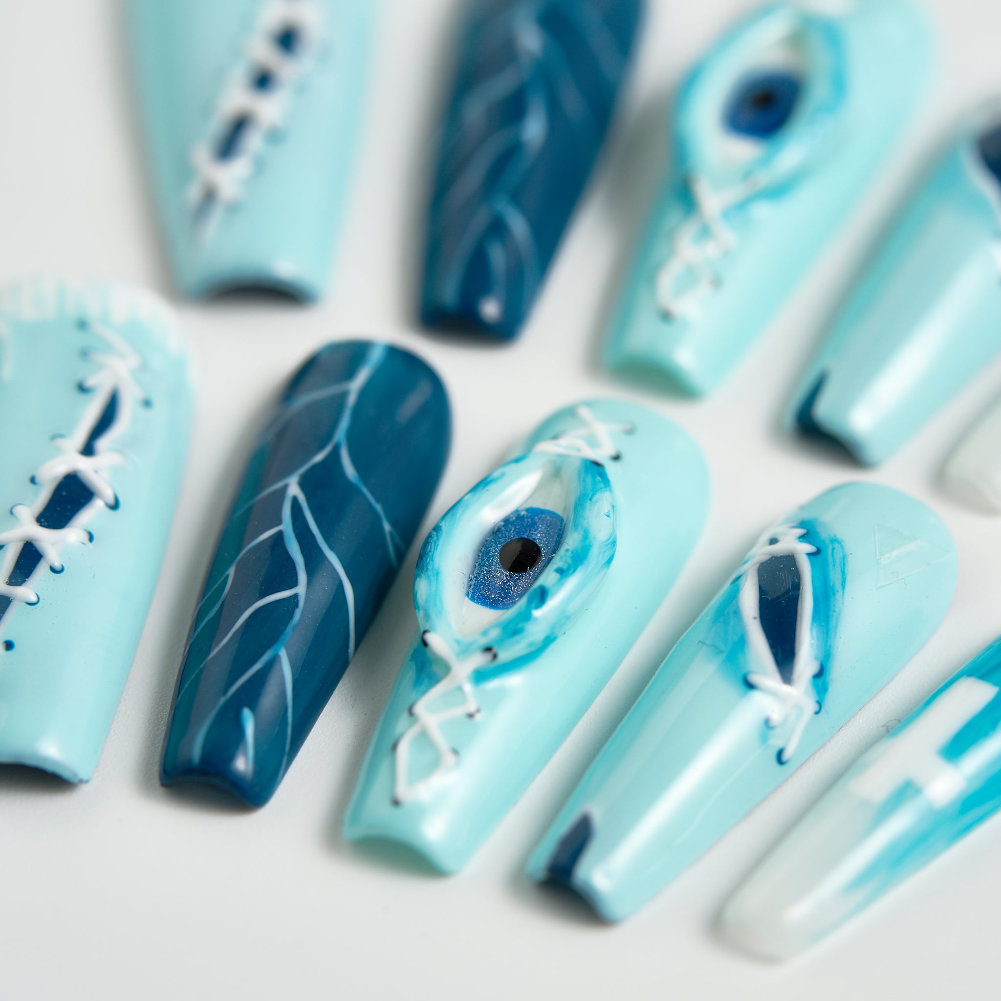Close-up of MiuNail Siren Hex press-on nails in icy blue and navy with lightning patterns, stitched accents, and a central eye motif, shown on a light background.