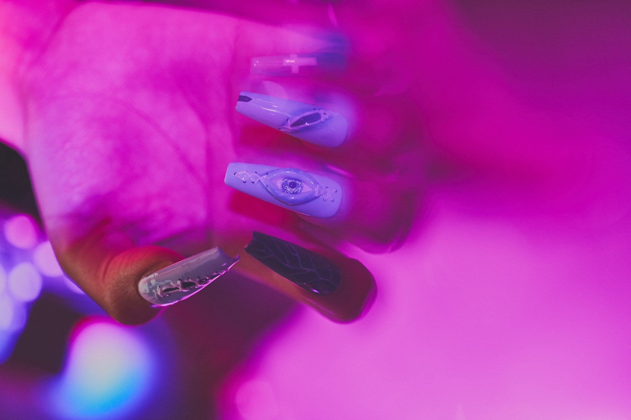 Hand with MiuNail Siren Hex press-on nails in icy blue and navy with stitched and eye motifs, shown under purple lighting in a night-time setting.
