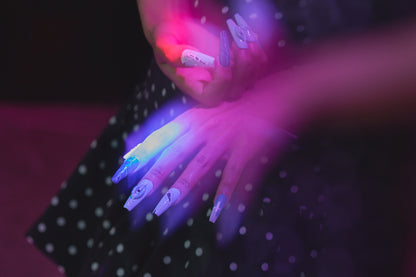 Hands wearing MiuNail Siren Hex press-on nails in icy blue and navy with stitched and lightning motifs, shown under purple and blue light in a night-time setting.