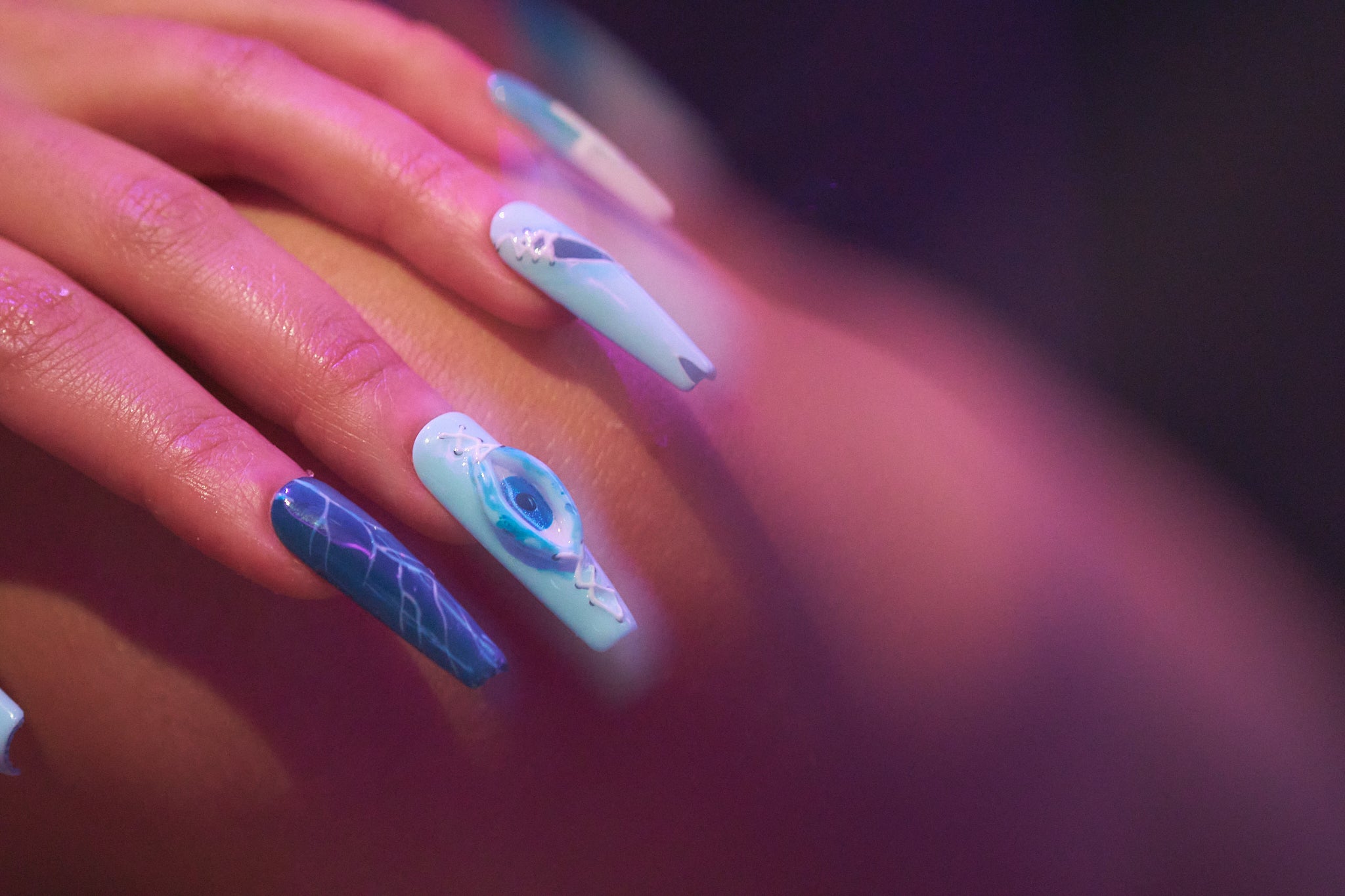 Close-up of MiuNail Siren Hex press-on nails in icy blue and navy with stitched and lightning motifs, shown under soft purple light on fingers at rest.