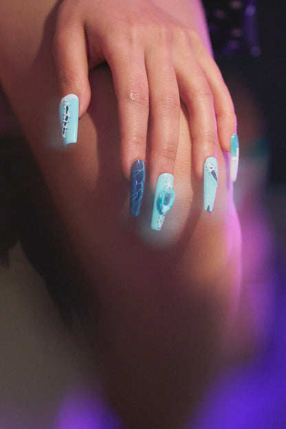 Hand wearing MiuNail Siren Hex press-on nails in icy blue and navy with lightning and stitched patterns, resting on thigh in soft purple light.