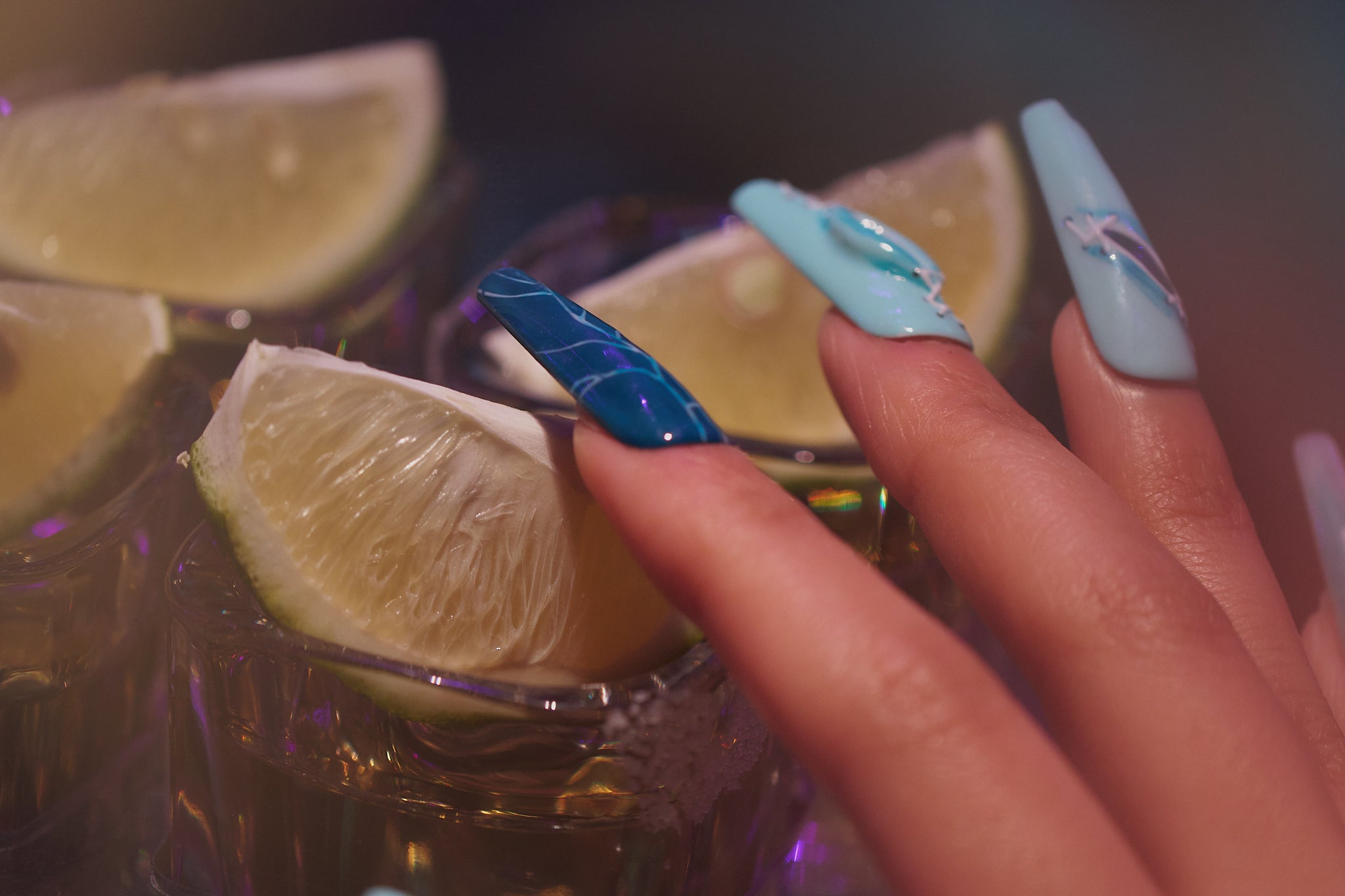 Hand with MiuNail Siren Hex press-on nails in blue and icy tones, touching a glass rim with lime wedges under party lighting.