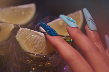 Hand with MiuNail Siren Hex press-on nails in blue and icy tones, touching a glass rim with lime wedges under party lighting.