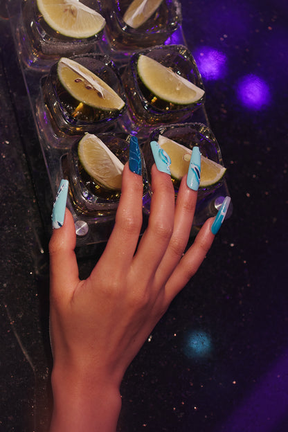 Hand with MiuNail Siren Hex press-on nails in icy blue tones reaching toward cocktail tray with lime wedges on a glittery black surface.