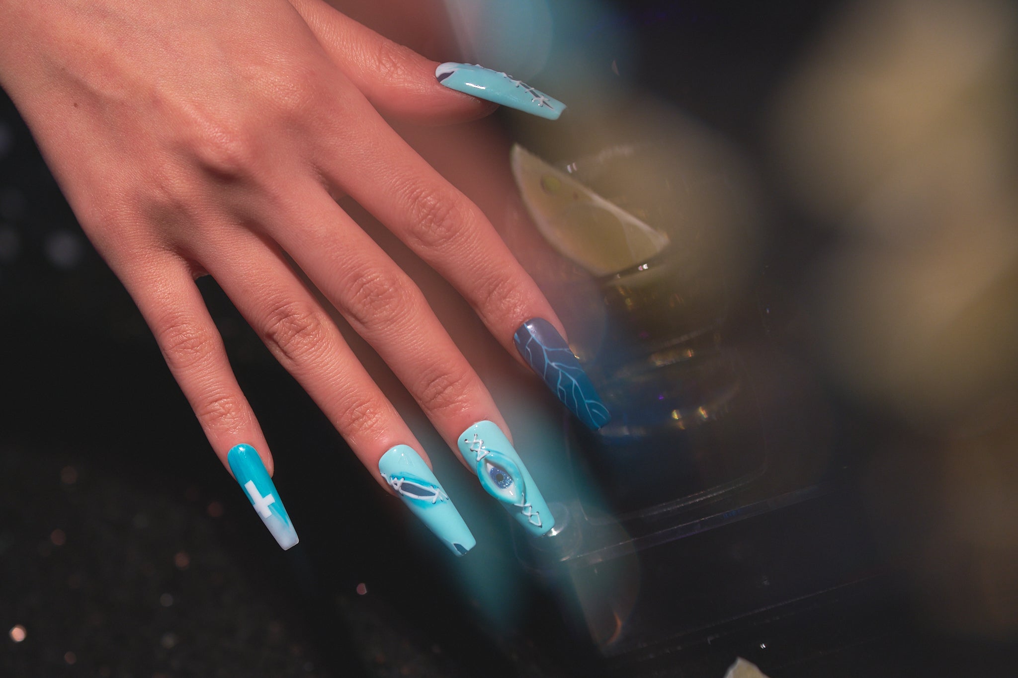 Hand wearing MiuNail Siren Hex press-on nails in icy blue and navy with lightning and stitched details, resting beside a cocktail glass in a dimly lit setting.