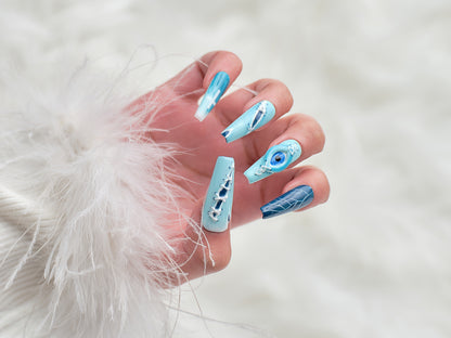 Hand wearing MiuNail Siren Hex press-on nails in icy blue and navy with stitched accents, lightning veining, and a central eye design, posed against a white faux fur background.