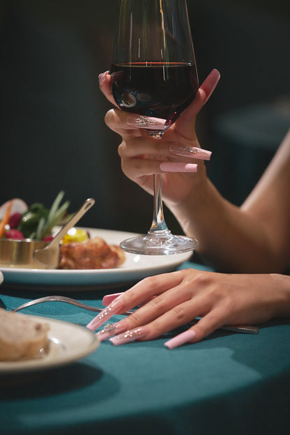 A graceful hand with glossy pastel pink nails holds a glass of red wine above a teal tablecloth, with elegant plating and soft background blur.
