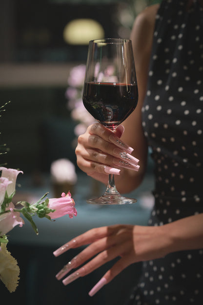 Woman in a polka dot dress holding a glass of red wine, showing Sugar Flutter pink press-on nails by MiuNail.