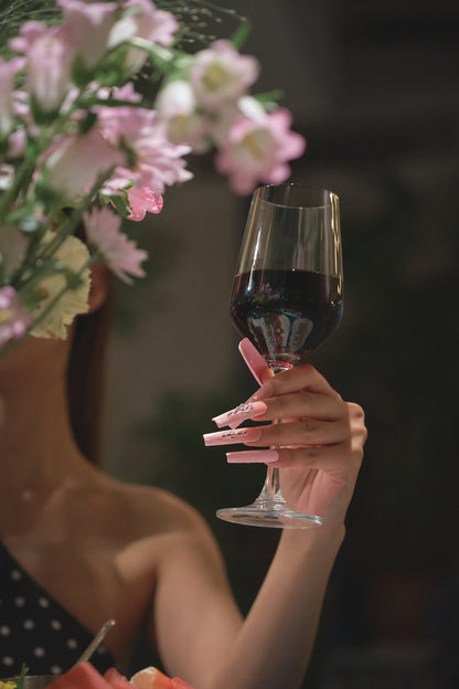 Hand with Sugar Flutter pink press-on nails holding a glass of red wine, blurred floral background adds an elegant touch.