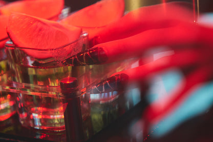 Hand with MiuNail Venom Twist black press-on nails reaching for a cocktail glass with citrus garnish, under moody red lighting.