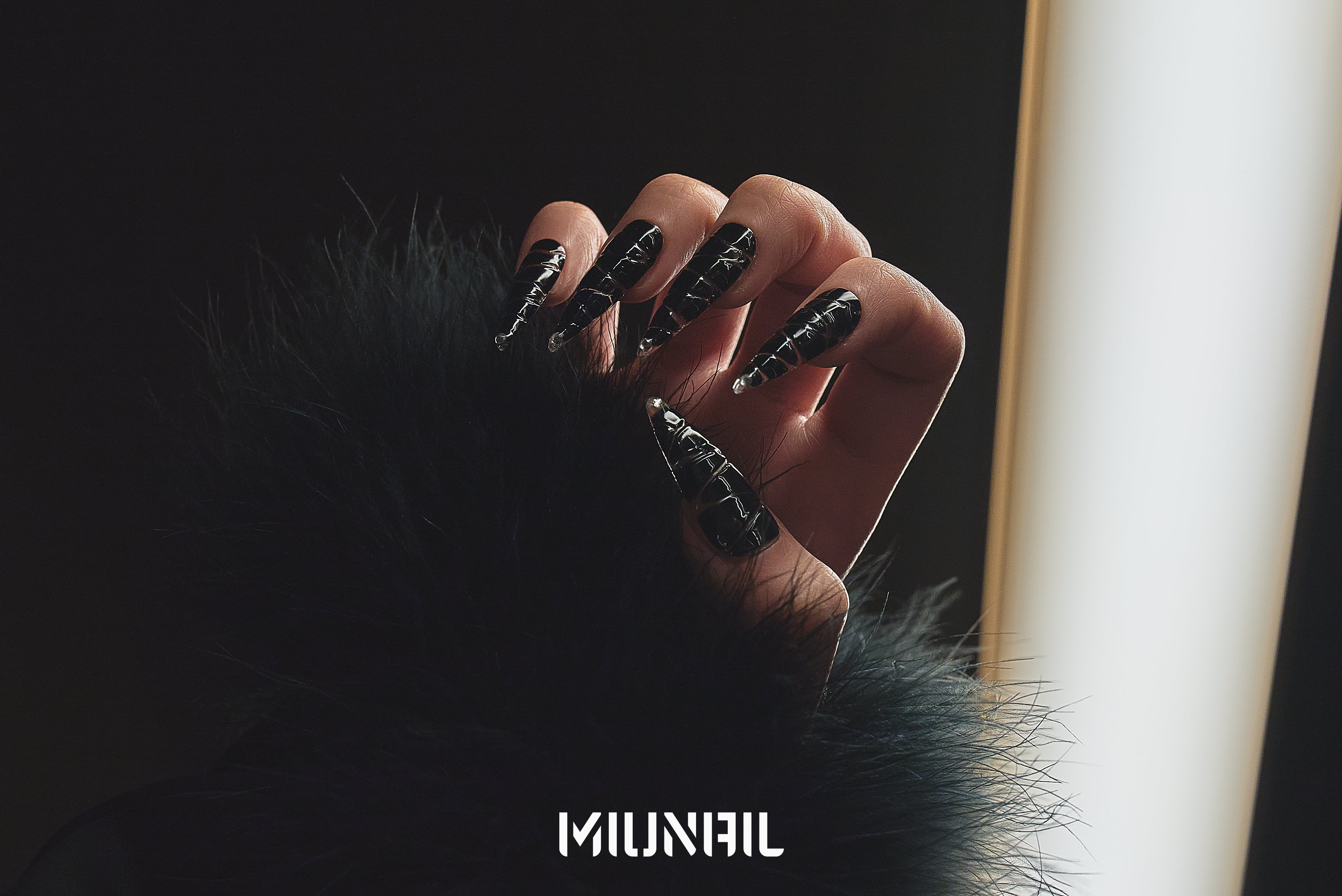 Close-up of a hand wearing MiuNail Venom Twist black press-on nails with metallic spiral accents, posed against a soft spotlight in a black faux fur coat.