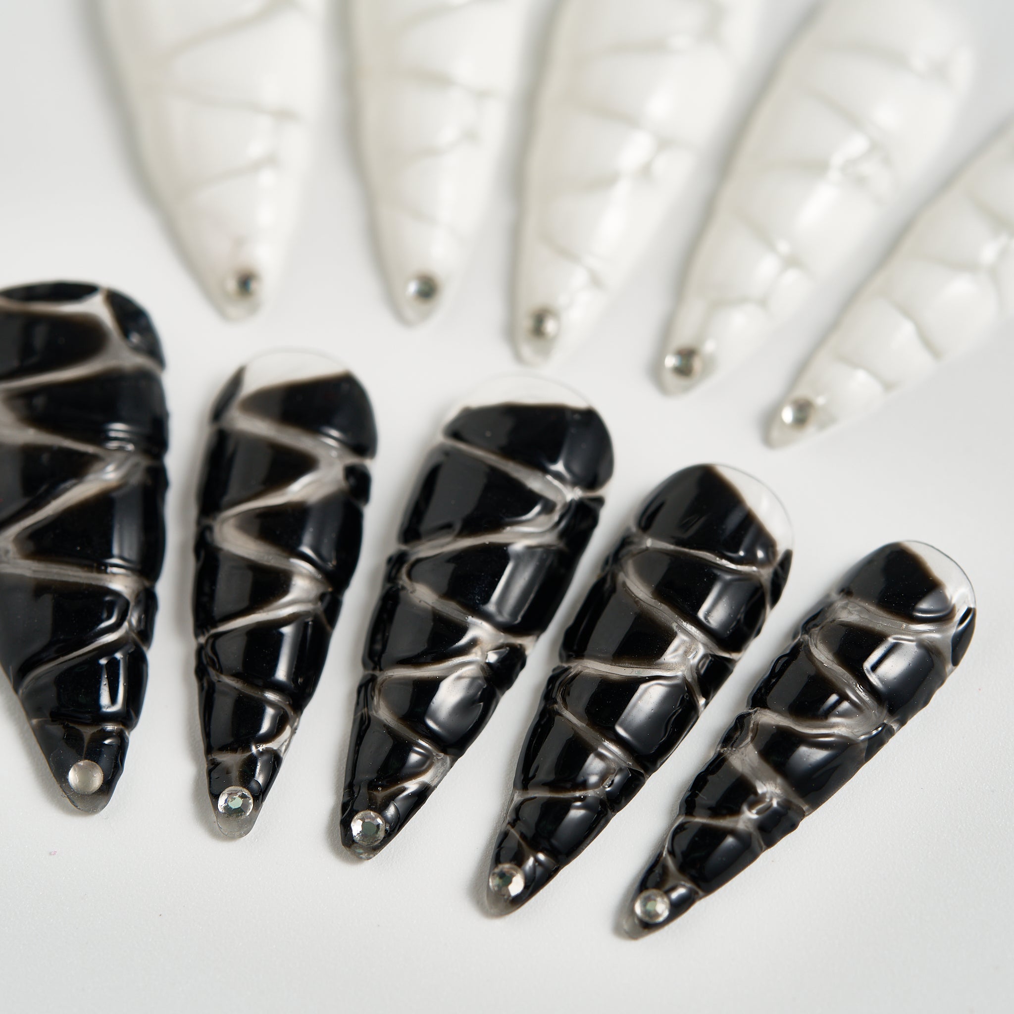 Macro view of MiuNail Venom Twist black press-on nails with glossy silver spiral wraps and rhinestone tips on a white background.