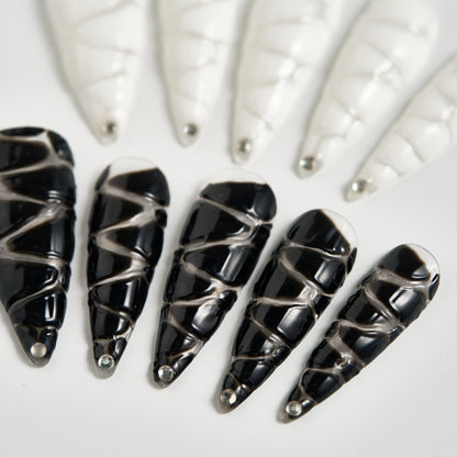Macro view of MiuNail Venom Twist black press-on nails with glossy silver spiral wraps and rhinestone tips on a white background.