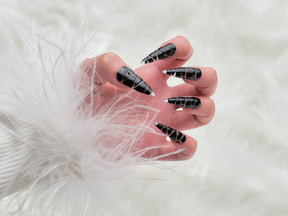 Hand wearing MiuNail Venom Twist black press-on nails with chrome spiral texture and pointed tips, shown against a soft white fur background.