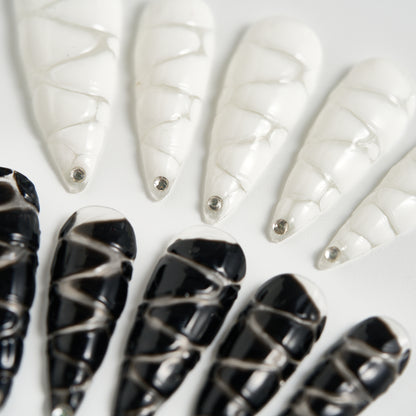 Close-up of MiuNail Venom Twist press-on nails in glossy black and white with silver spiral detailing, displayed on a white background.