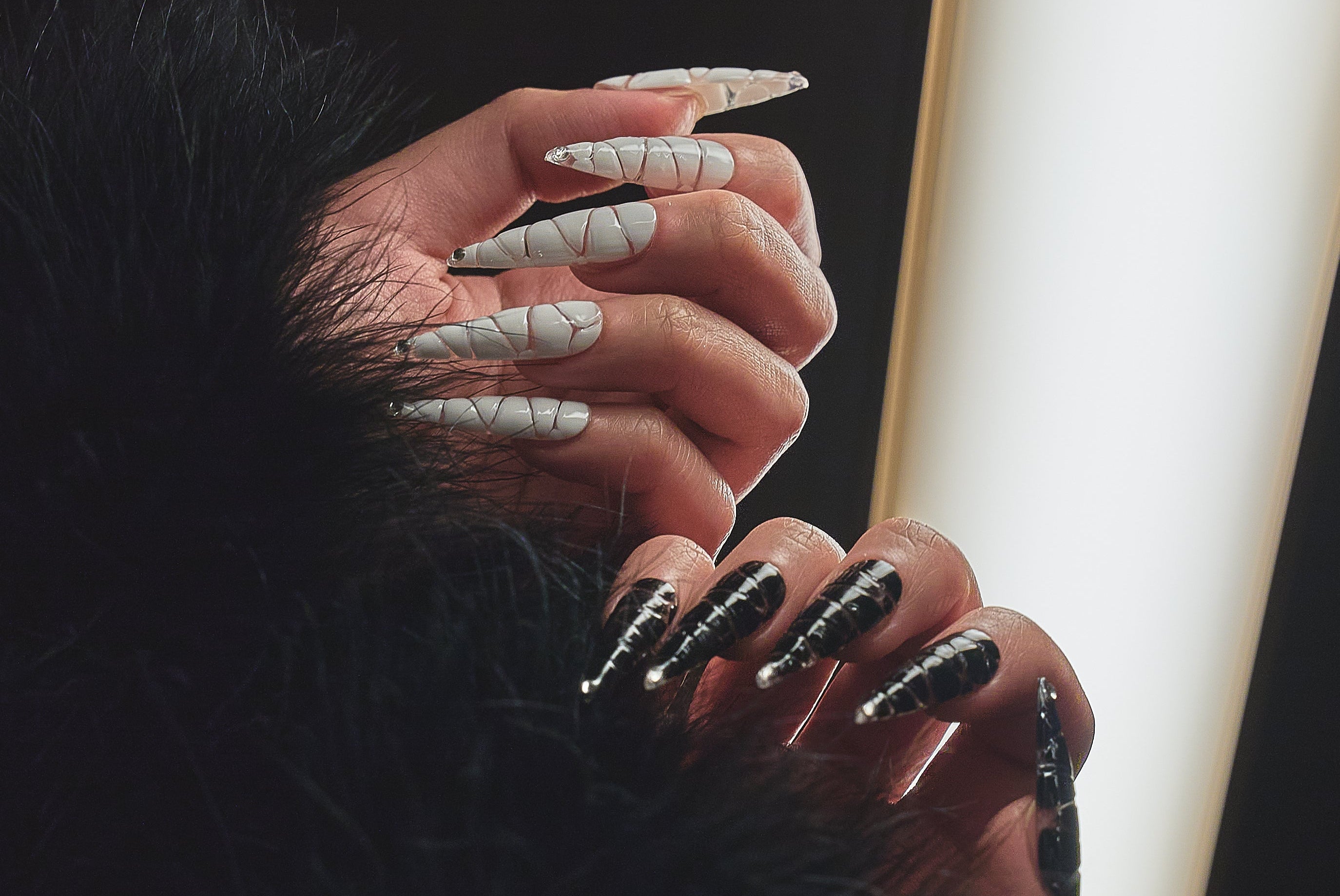 Close-up of two hands wearing MiuNail Venom Twist black and white press-on nails with metallic spirals, styled with black faux fur and soft backlight.